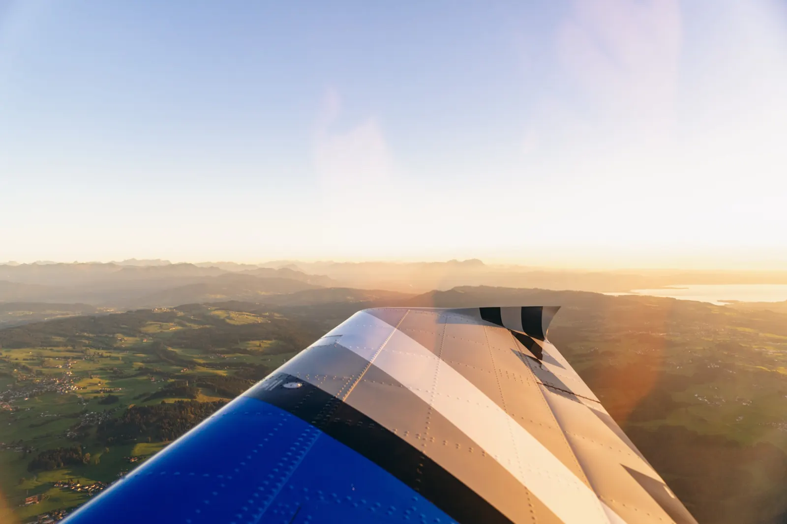 Blick über die blaue Tragfläche auf eine Hügellandschaft, am Horizont die Alpen