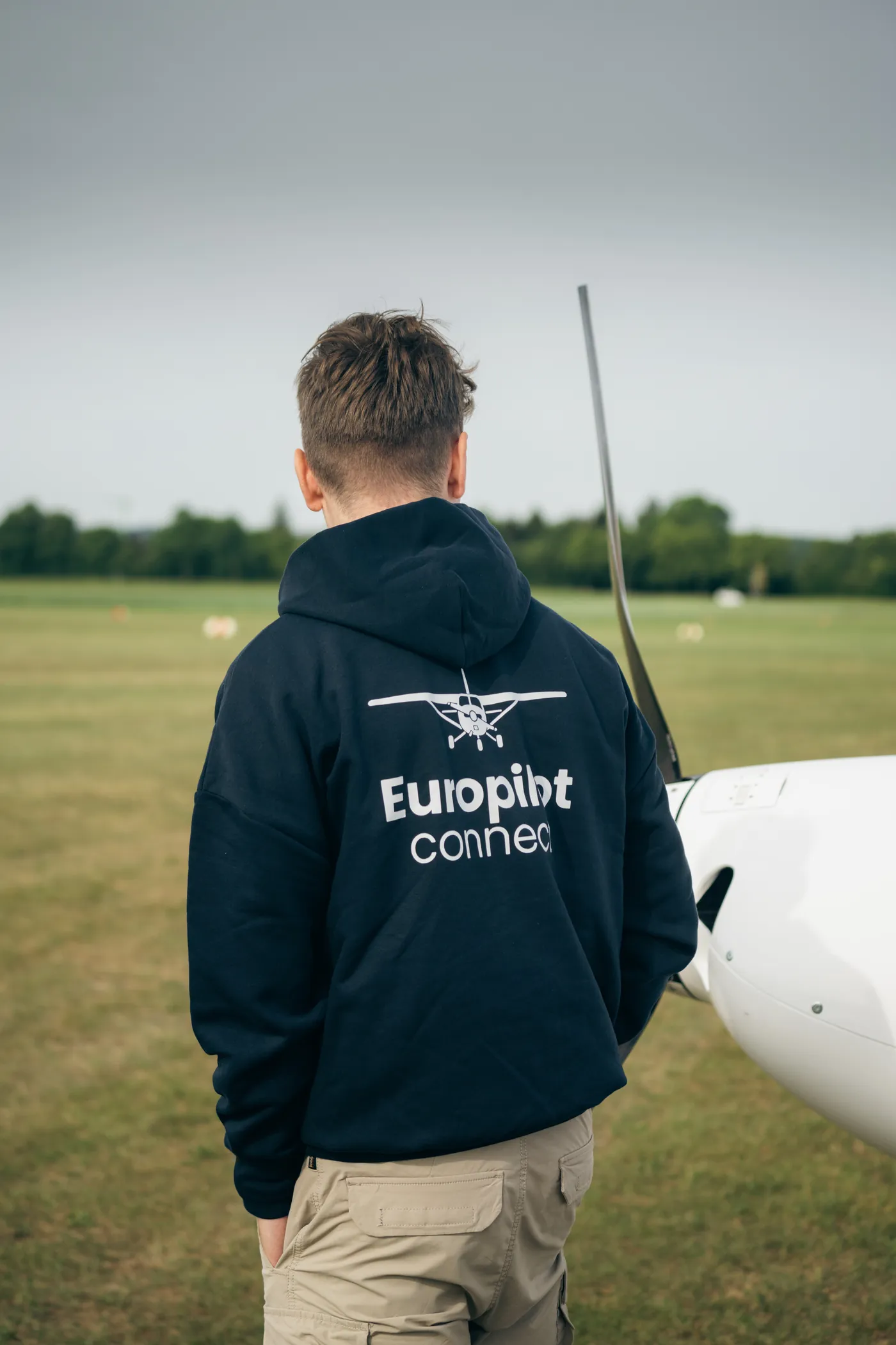 Pilot im EuroPilot-Connect-Hoodie geht über die Graspiste zu einem Flugzeug